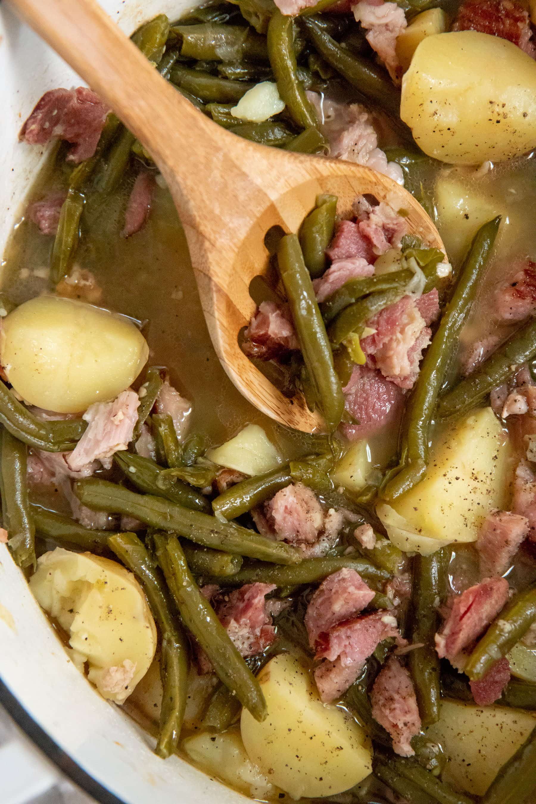 Close up of a wooden spoon in a pot of beans, ham, and new potatoes