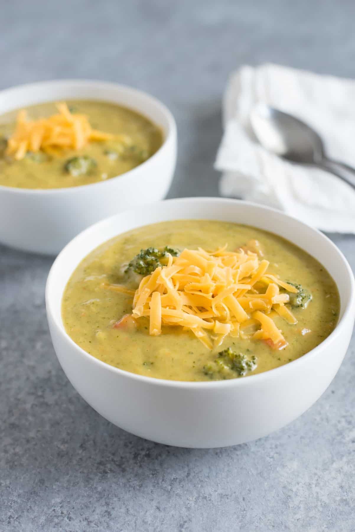 Two servings of broccoli cheddar and potato soup in white bowls garnished with shredded cheddar.