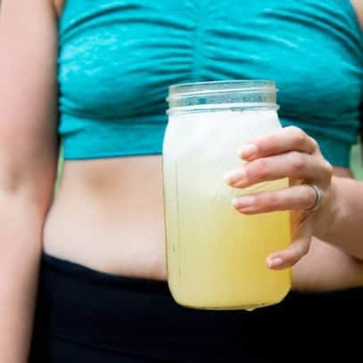 Torso of a woman in a turquoise sports bra and black pants, holding a glass jar of Homemade All-Natural Electrolyte Drink