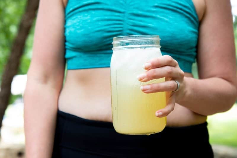 Torso of a woman in a turquoise sports bra and black pants, holding a glass jar of Homemade All-Natural Electrolyte Drink
