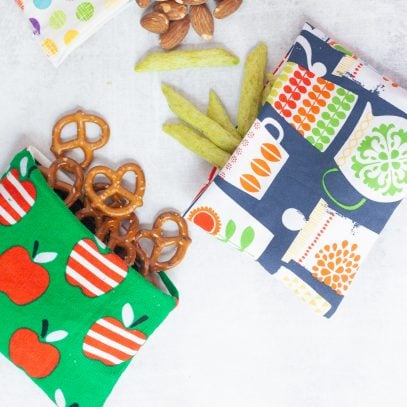 Three reusable snack bags, each spilling out a different snack - pretzels, almonds, and crispy snap peas