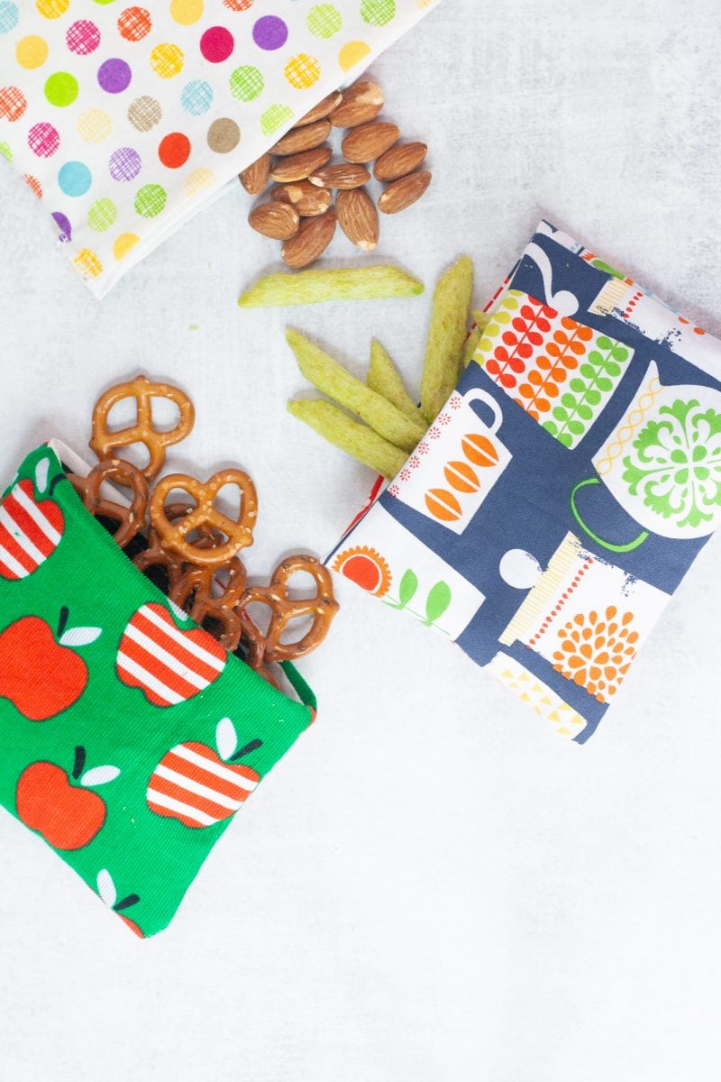 Three reusable snack bags, each spilling out a different snack - pretzels, almonds, and crispy snap peas