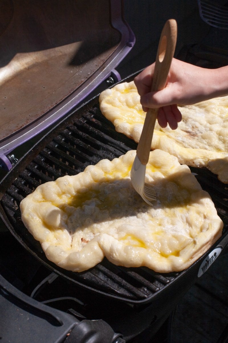Side angle shot of pizza dough on a grill grate being brushed with oil
