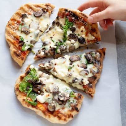 Overhead shot of a hand grabbing a slice of Grilled Flatbread Pizza with marinara, mushrooms, spinach, and goat cheese