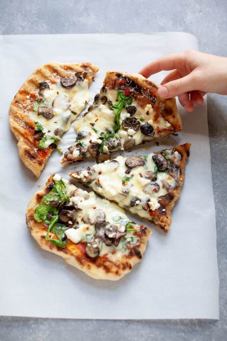 Overhead shot of a hand grabbing a slice of Grilled Flatbread Pizza with marinara, mushrooms, spinach, and goat cheese