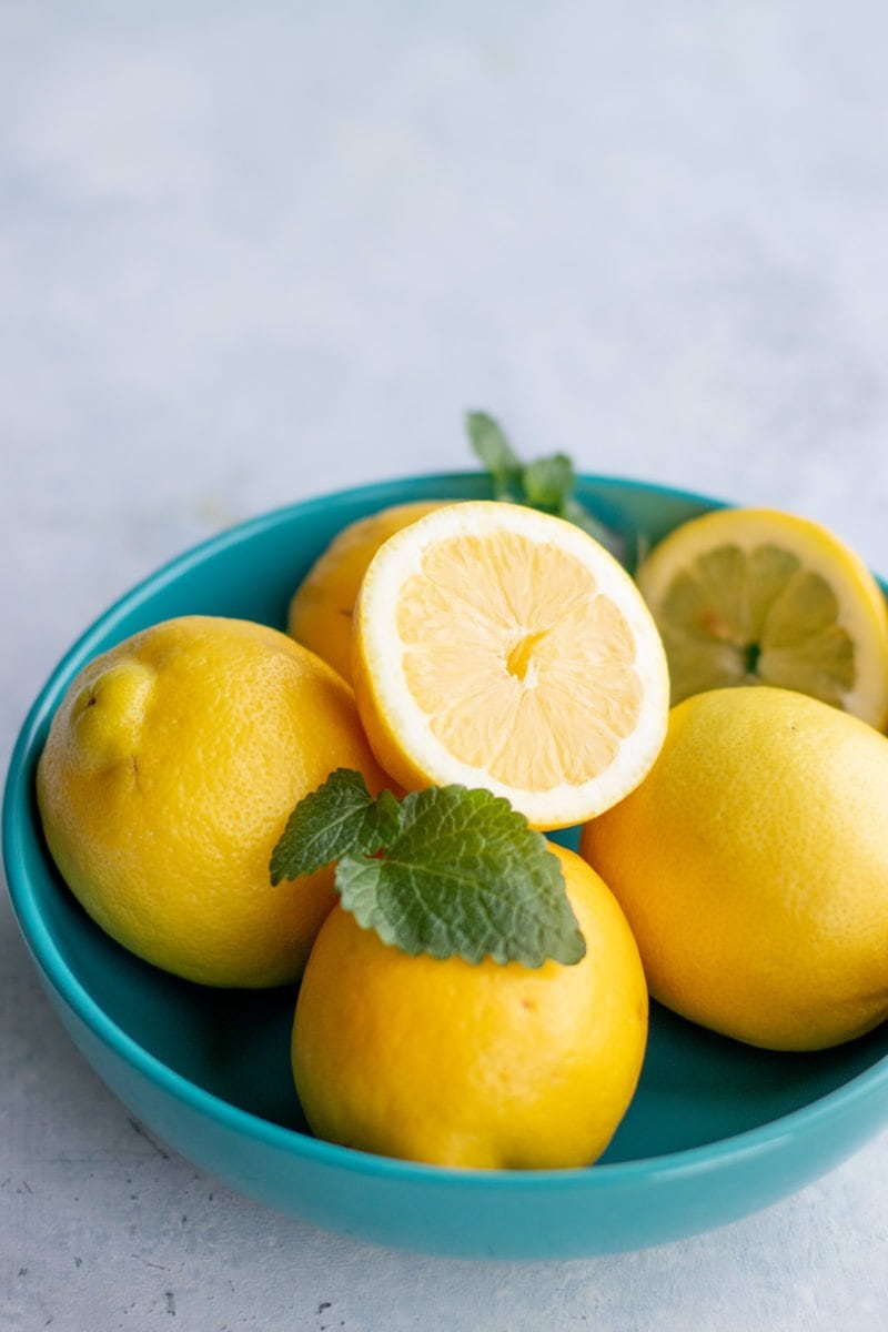 Side angle shot of whole and halved lemons in a blue bowl on a light background