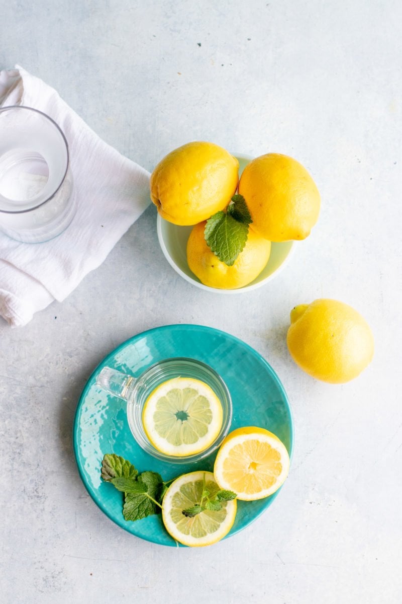 Overhead shot of a clear mug of lemon water on a blue plate, with a bowl of whole lemons nearby