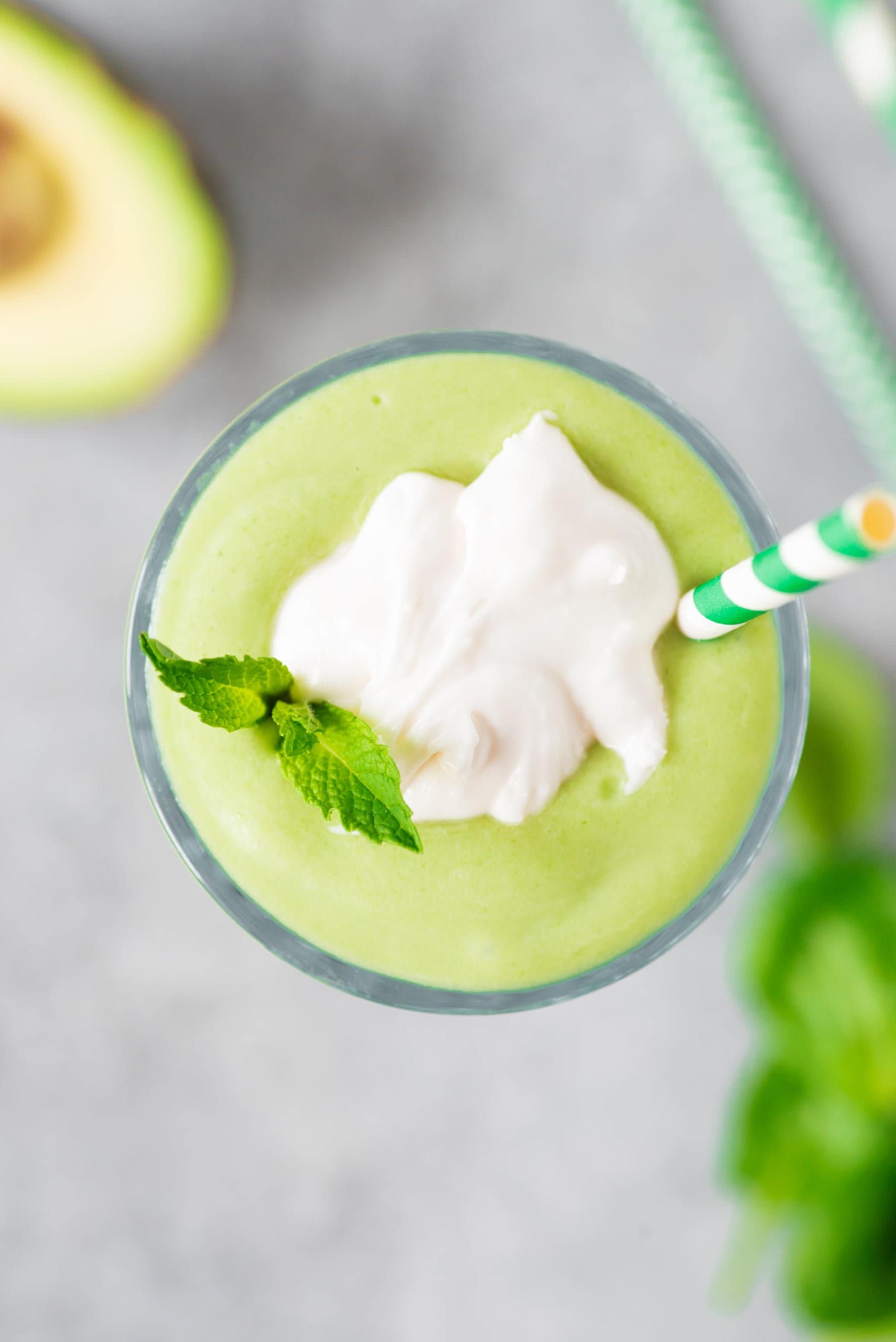 A creamy avocado banana smoothie garnished with coconut whipped cream, fresh mint leaves, and a striped straw.