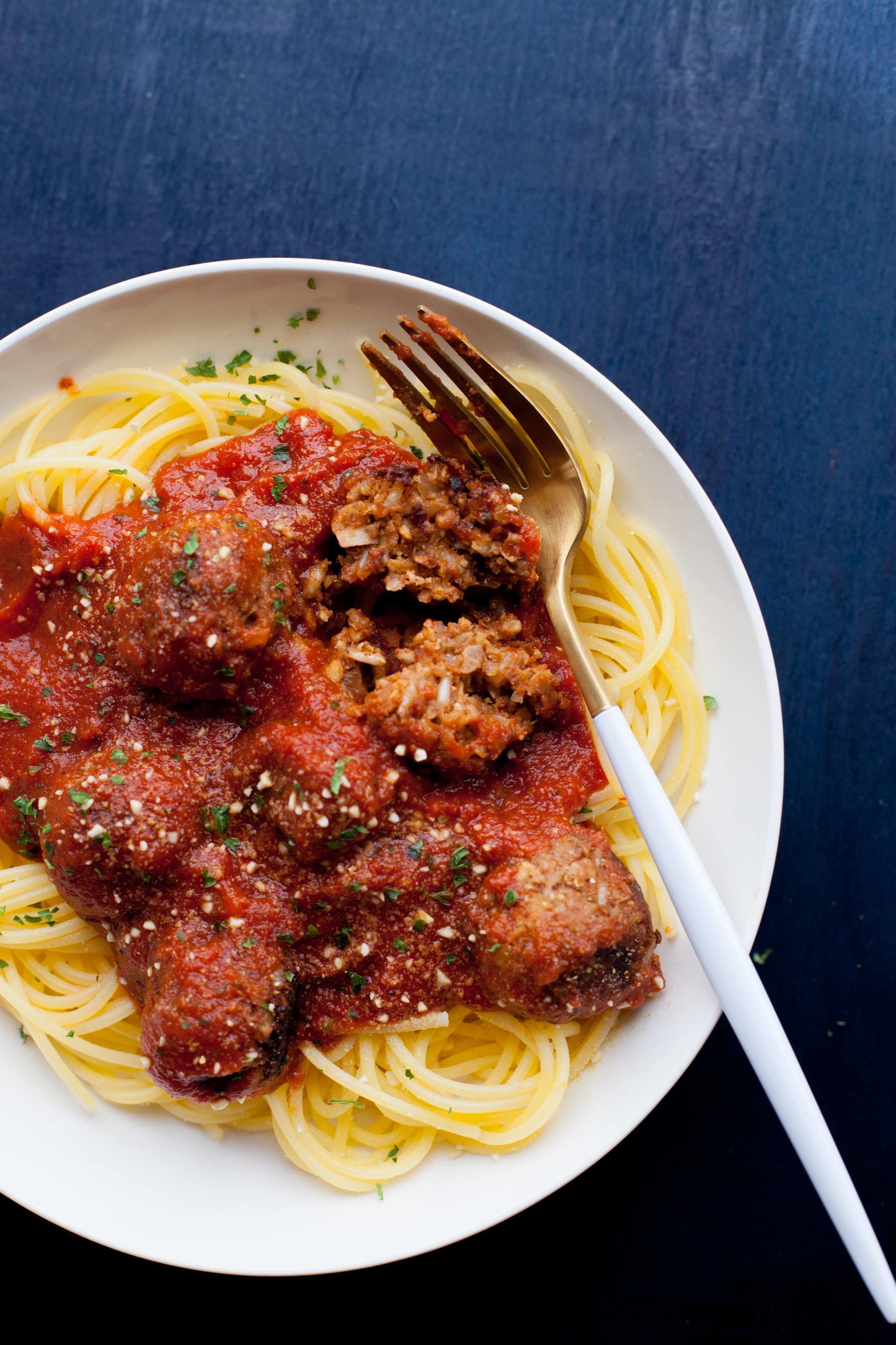 White plate filled with spaghetti, vegan meatballs, and marinara sauce on a blue background. A white-handled fork rests on the edge of the plate.