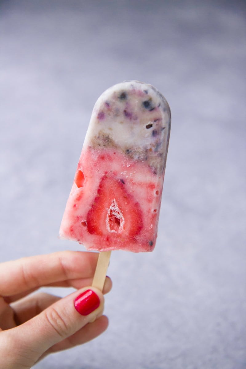 A hand holds up a red, white, and blue berry yogurt popsicle