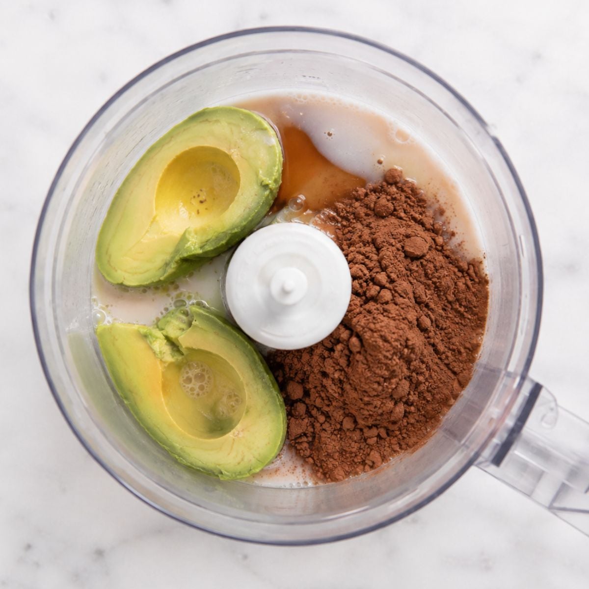 Halved avocado, almond milk, and cocoa powder in a food processor before blending into avocado chocolate pudding.