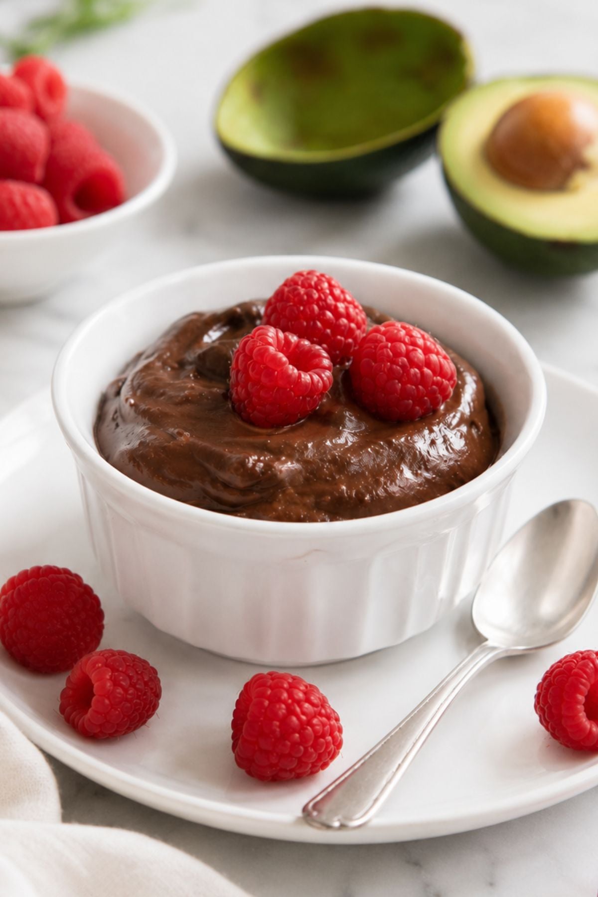 A bowl of avocado chocolate pudding topped with fresh raspberries, served with a spoon and a halved avocado in the background.