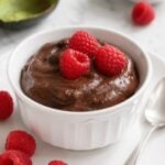 An avocado and chocolate dessert in a ramekin, topped with three raspberries and served with a silver spoon.