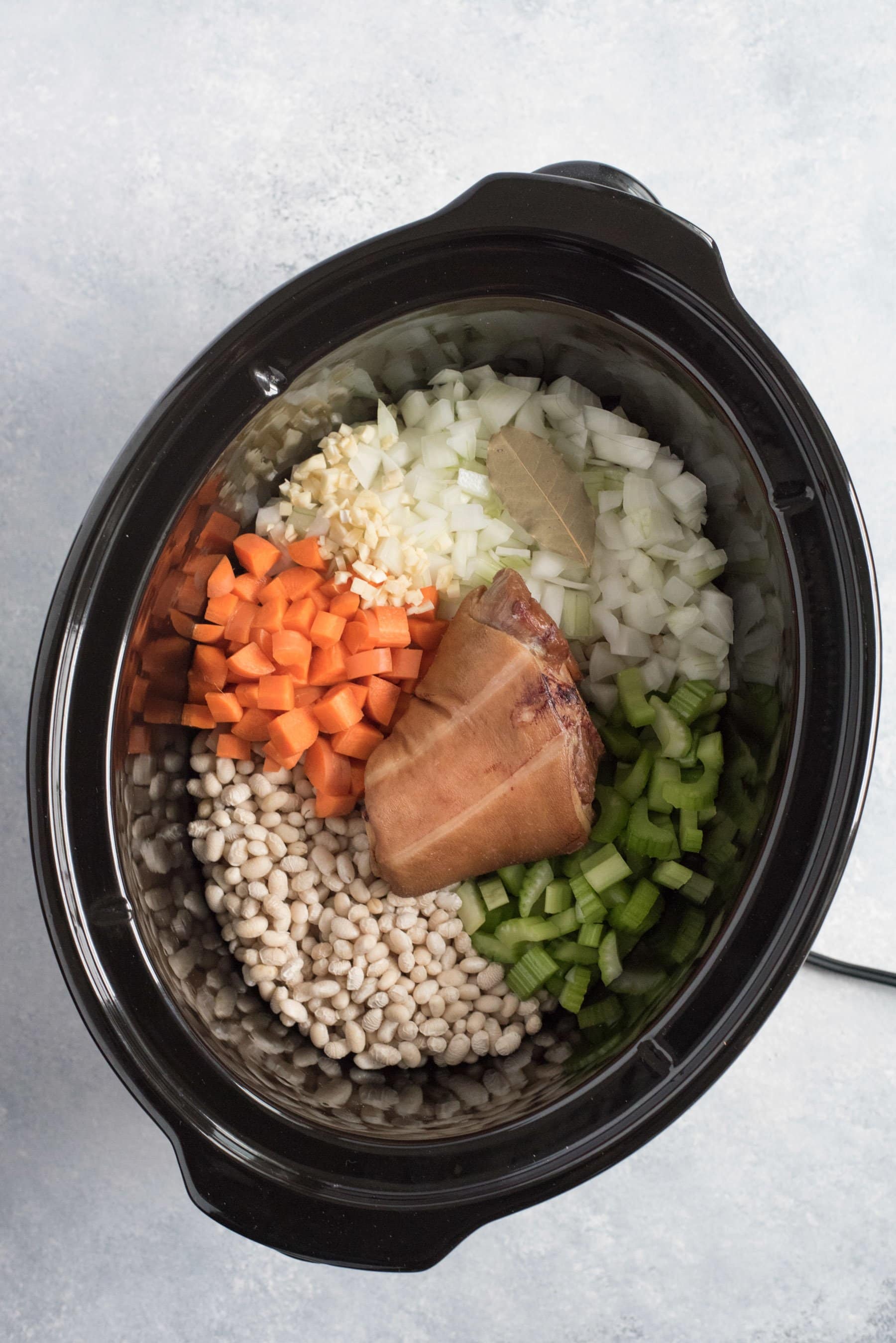 Carrots, onion, bay leaves, celery, dried beans, and a ham hock sit in the black crock of a slow cooker.
