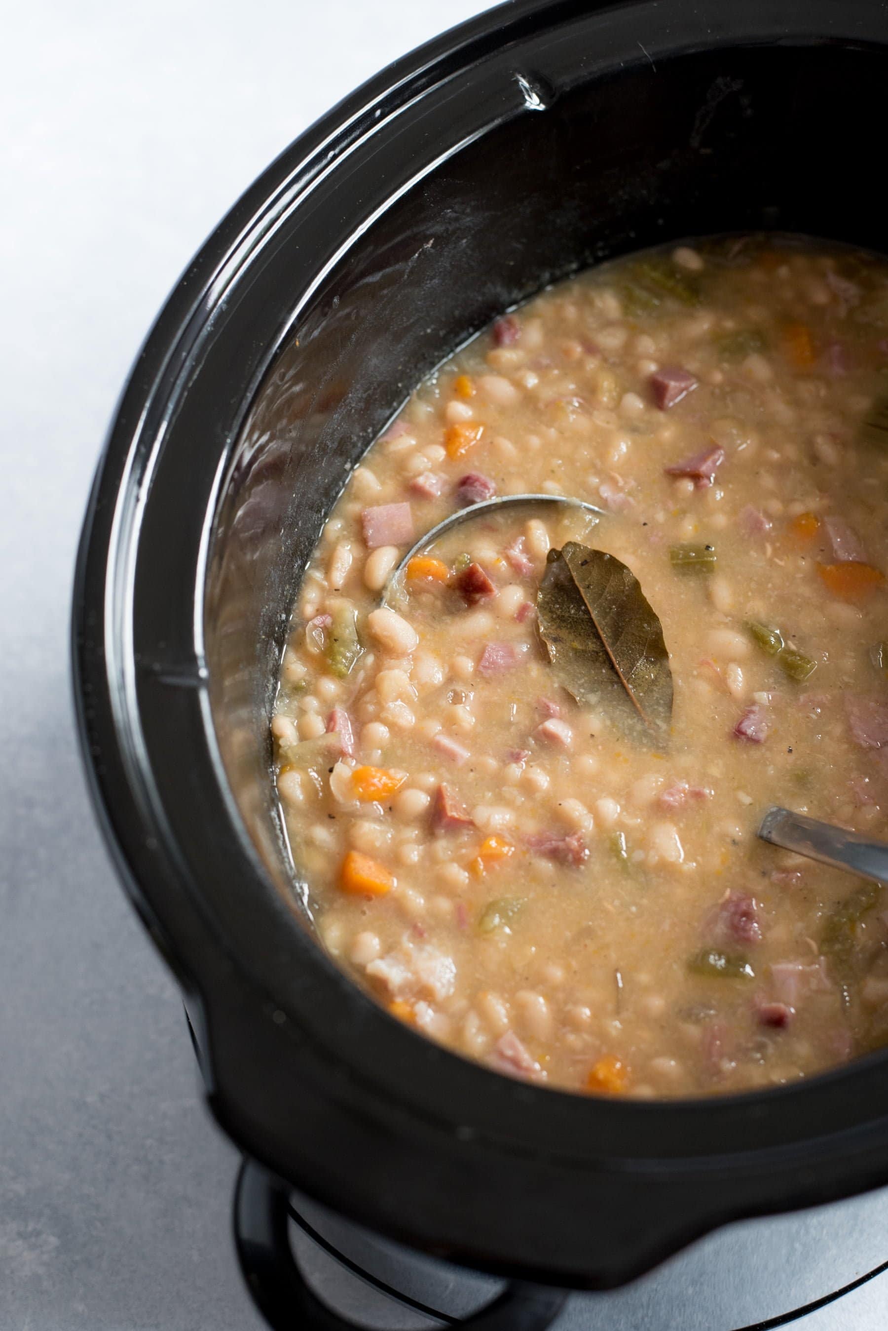 Bean soup cooks inside of a black slow cooker. A Bay leaf is seen floating on top, as a ladle dips into the pot.