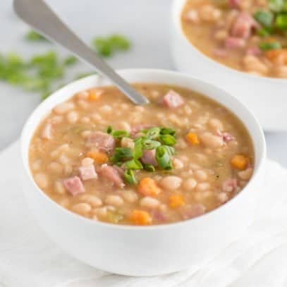 Cooked ham and beans in a white bowl with a spoon and scallion garnish.
