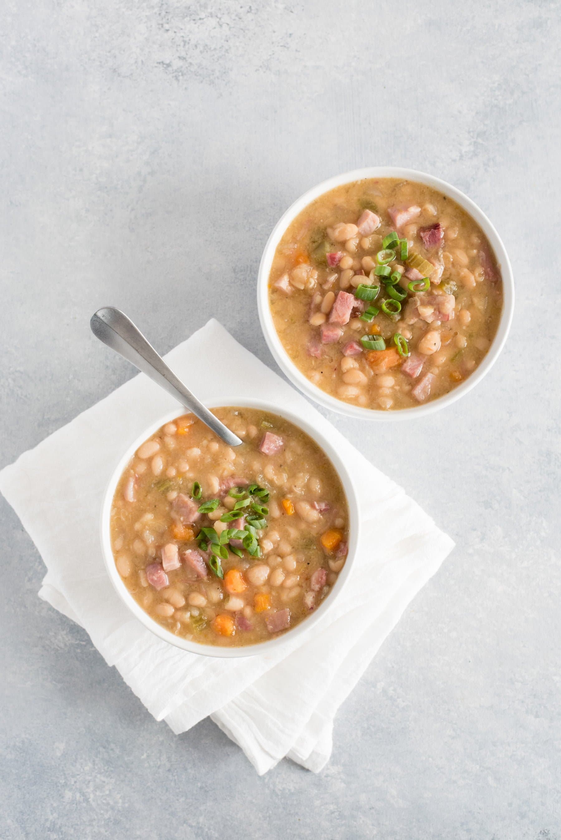 Two bowls of ham and bean soup, one with a spoon in it, sit on a counter.