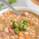 Cooked ham and beans in a white bowl with a spoon and scallion garnish. A text overlay reads Slow Cooker Ham and Beans.