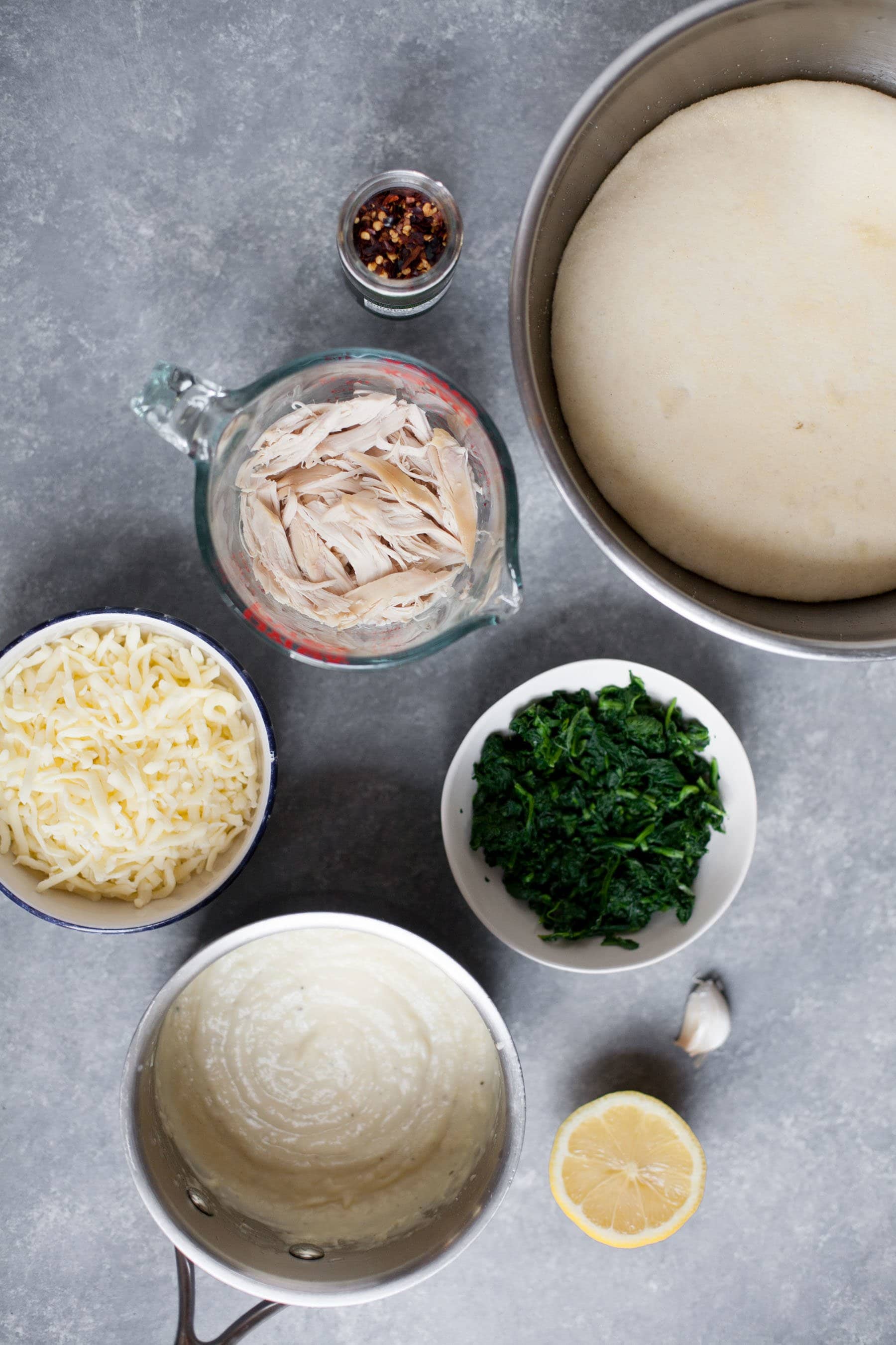 Pizza ingredients sit in bowls together - chicken, spinach, alfredo sauce, cheese, pizza dough