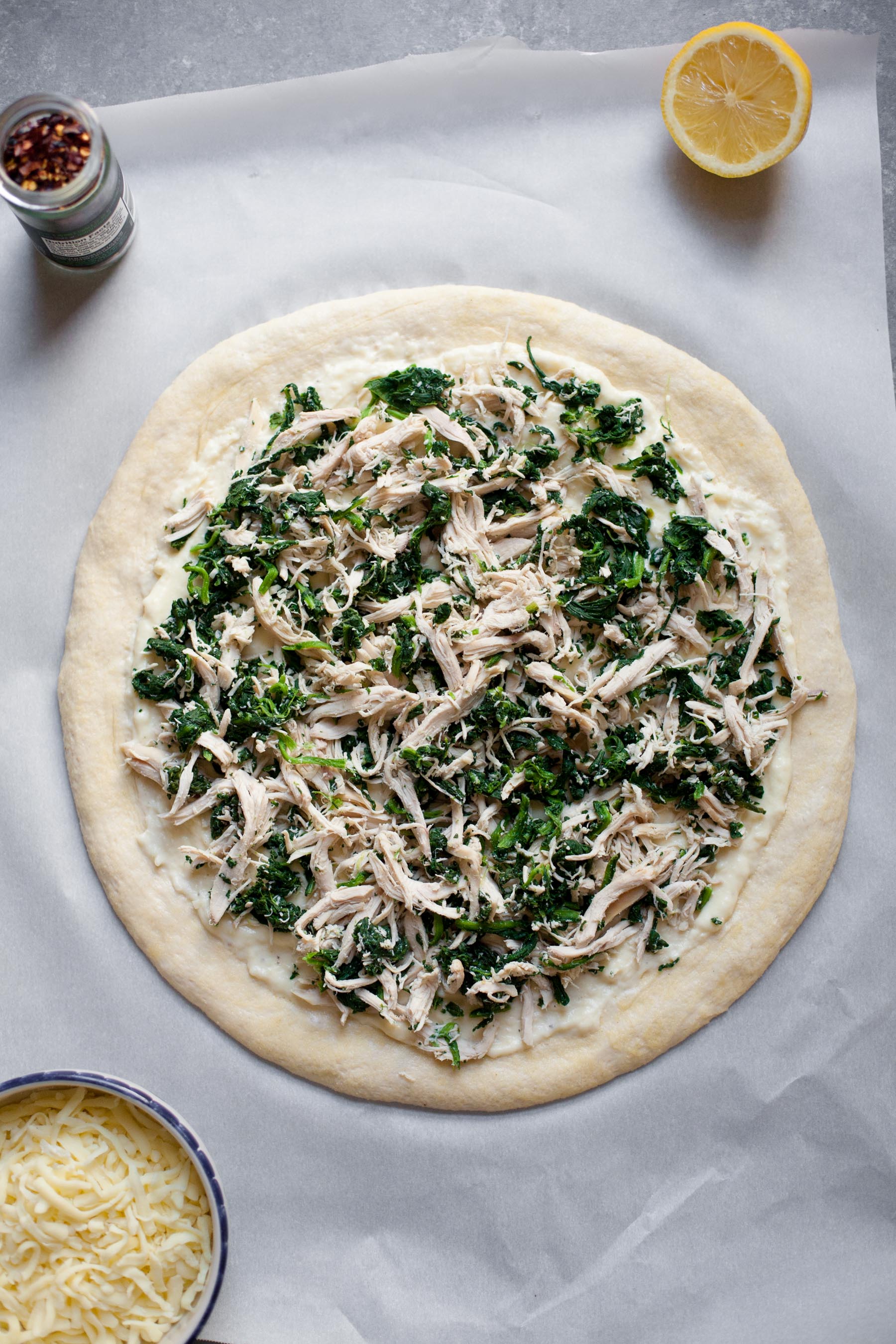 Uncooked Chicken Spinach Alfredo Pizza sits on a white background. A lemon and some spices sit nearby