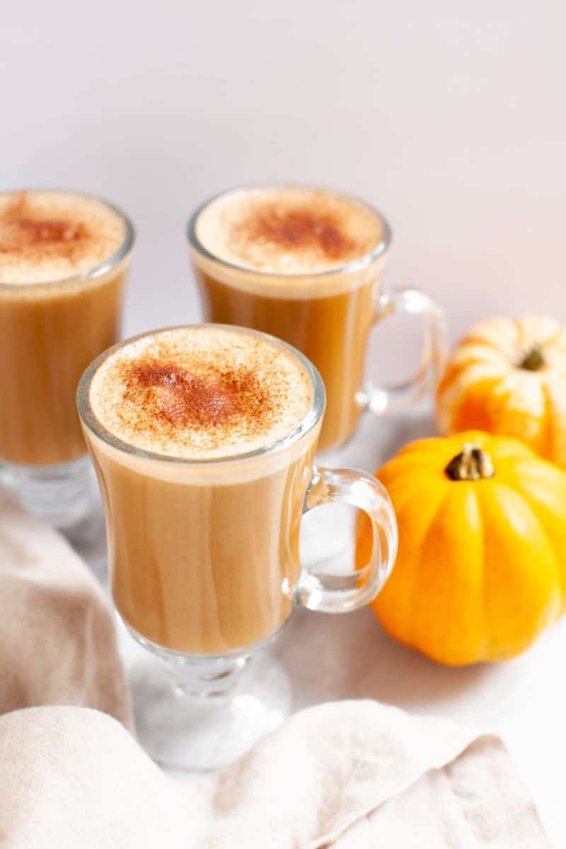 Three glass mugs filled with pumpkin spice lattes and topped with cinnamon, with mini pumpkins nearby.