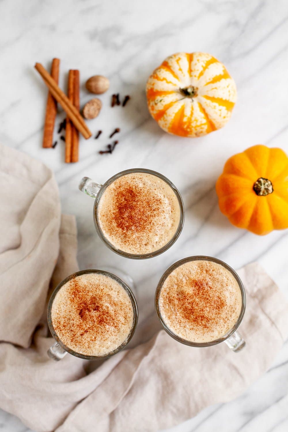 Overhead of three glass mugs filled with lattes and topped with cinnamon, with whole spices and mini pumpkins nearby.