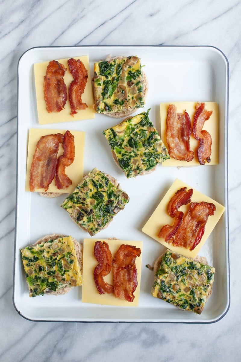 Overhead shot of open meal prep breakfast sandwiches with egg patties, cheese, and bacon on English muffins