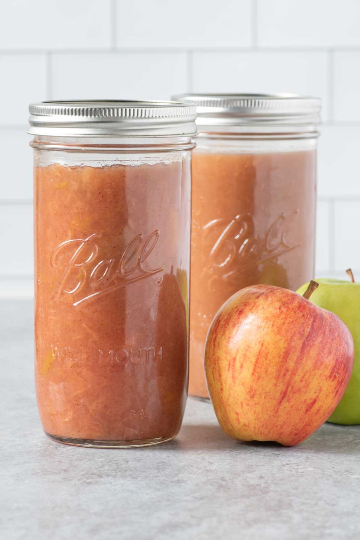 Two jars of applesauce sit on a gray countertop with apples beside them