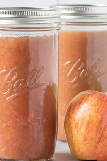 Two jars of applesauce sit on a gray countertop with apples beside them