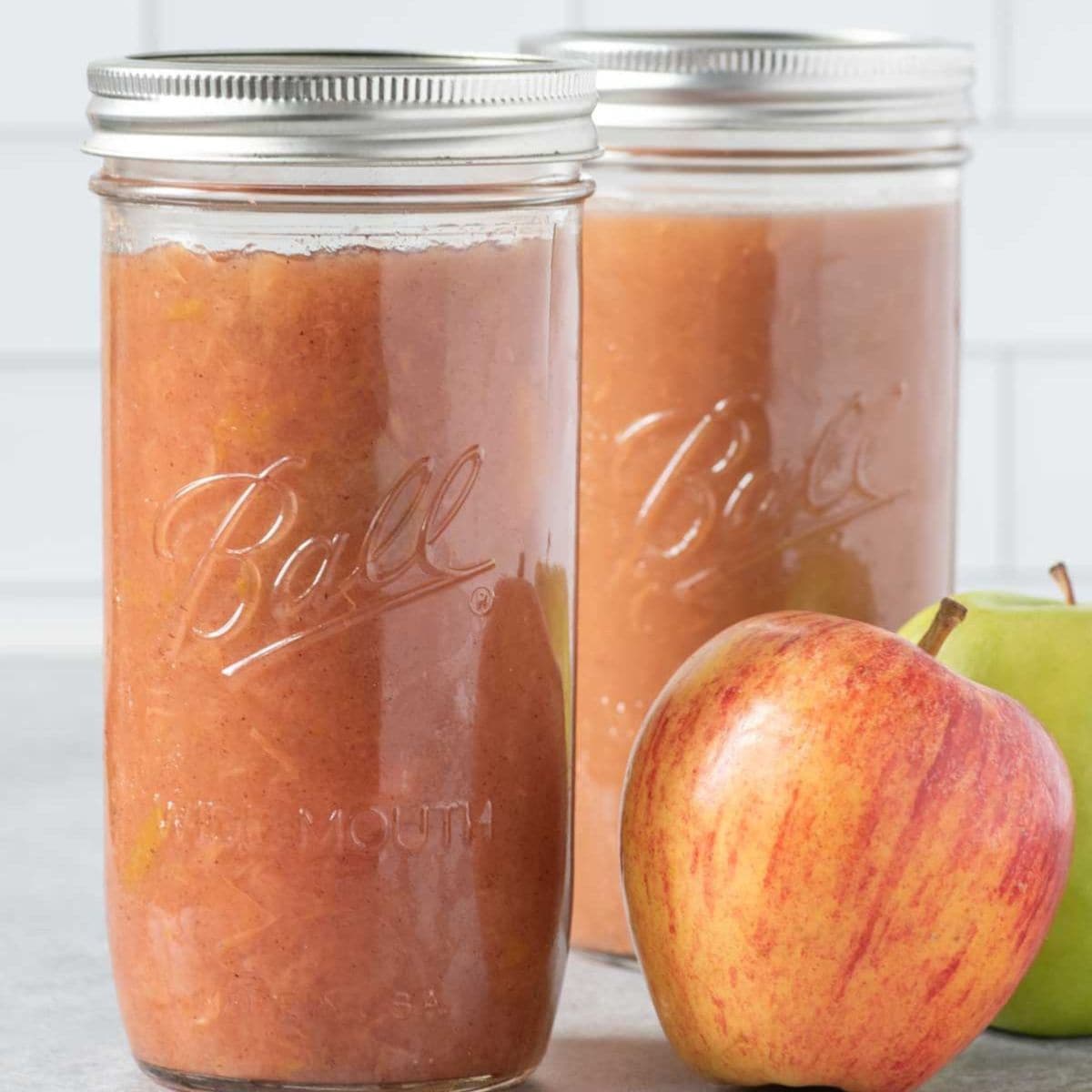 Two jars of applesauce sit on a gray countertop with apples beside them