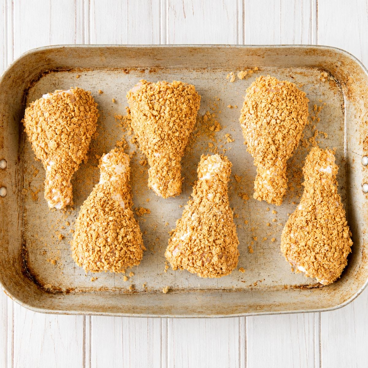 Six breaded chicken drumsticks arranged on a baking sheet before baking