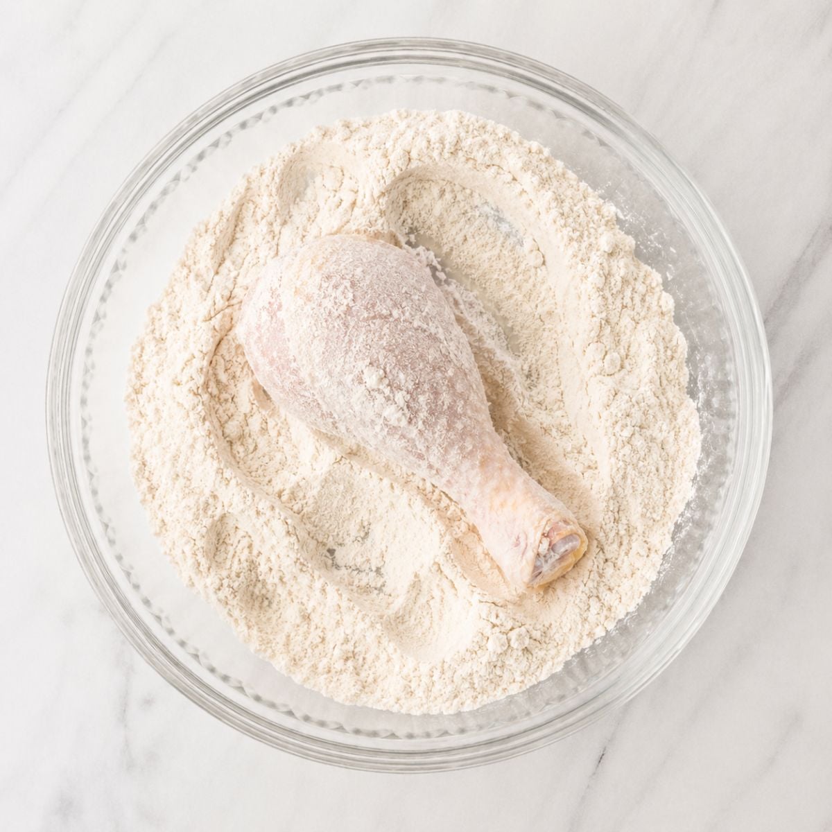 Raw chicken drumstick coated in flour in a glass bowl