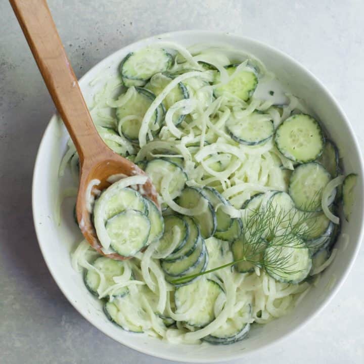 Overhead of dressed cucumber onion salad in a bow with a wooden spoon and fresh dill garnish.