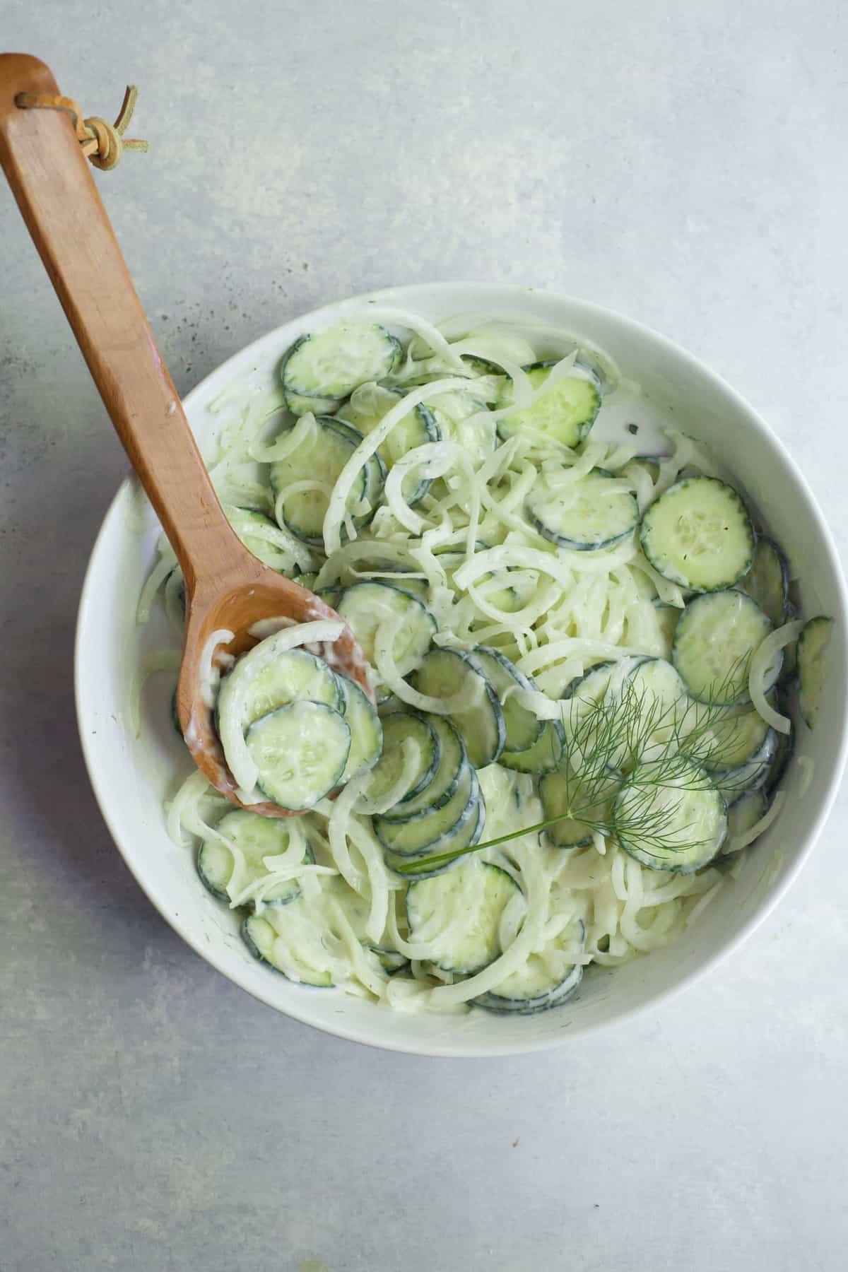 Overhead of dressed cucumber onion salad in a bow with a wooden spoon and fresh dill garnish.