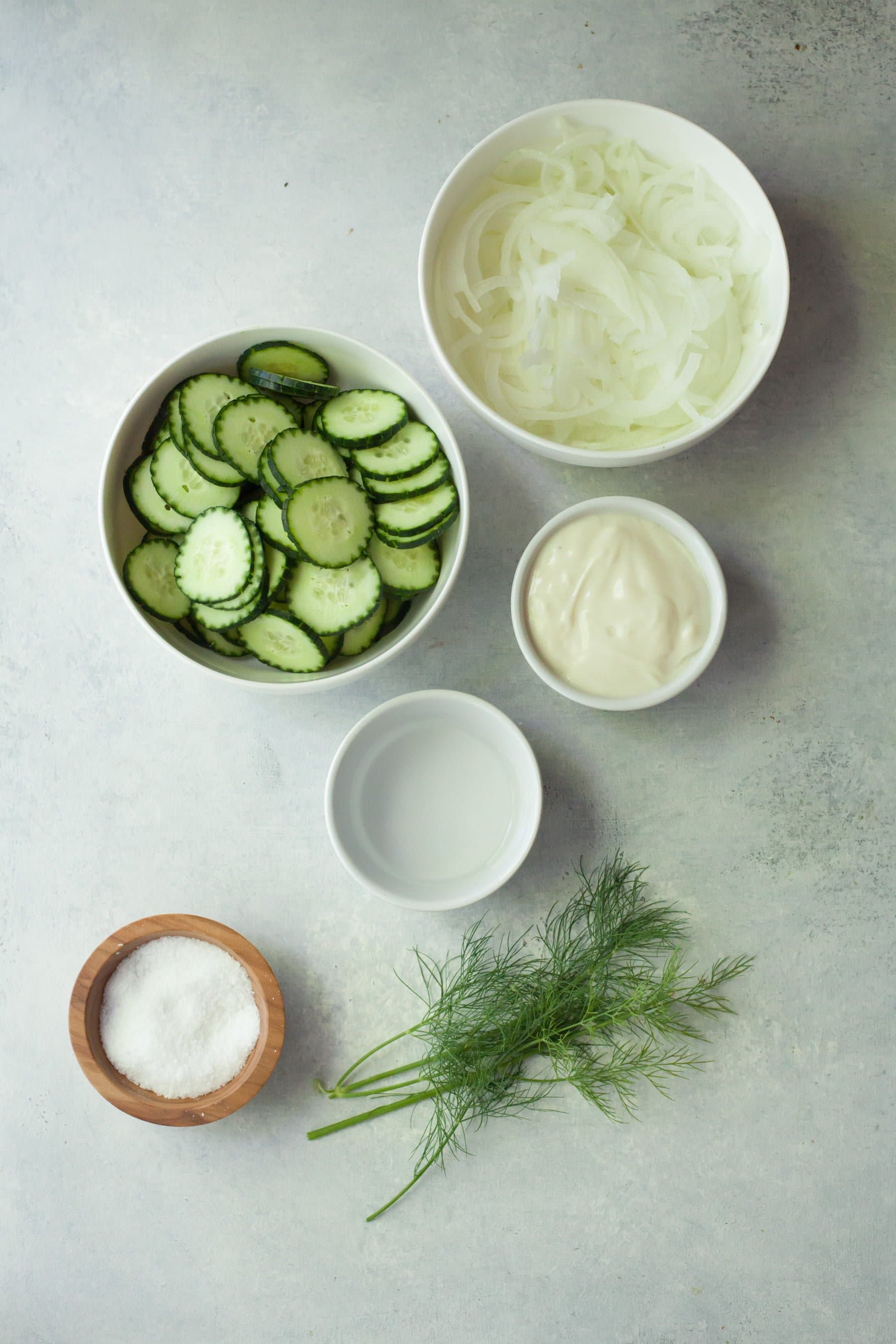 Overhead of six ingredients needed to make cucumber onion salad in bowls on a counter.