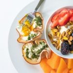 Egg cups made with turkey and herbs sit on a white plate. A bowl of fruit and yogurt sits to the side.