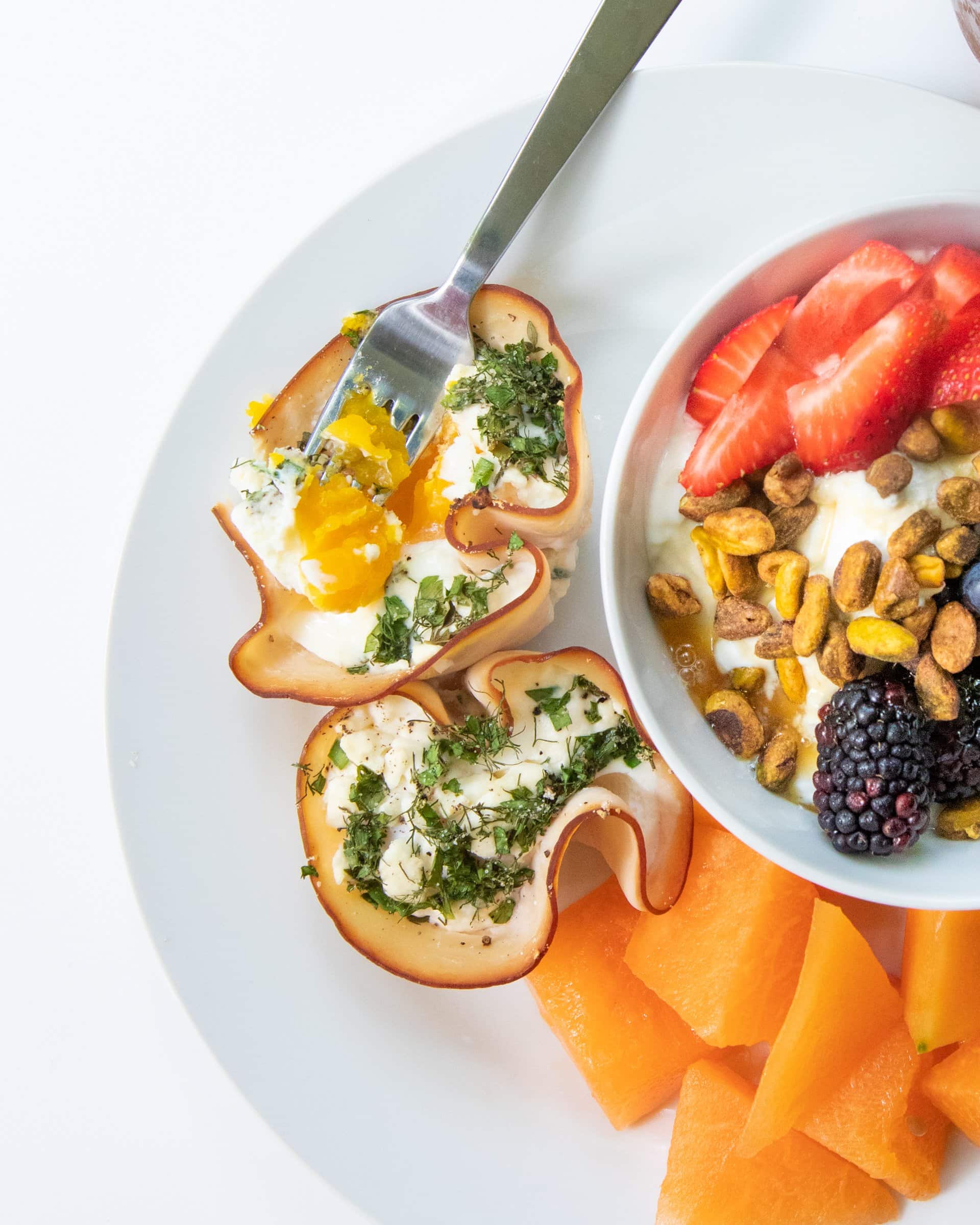 Egg cups made with turkey and herbs sit on a white plate. A bowl of fruit and yogurt sits to the side.