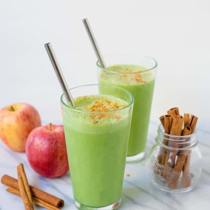 Two glasses of bright green smoothie with straws on white background with apples and cinnamon sticks garnish