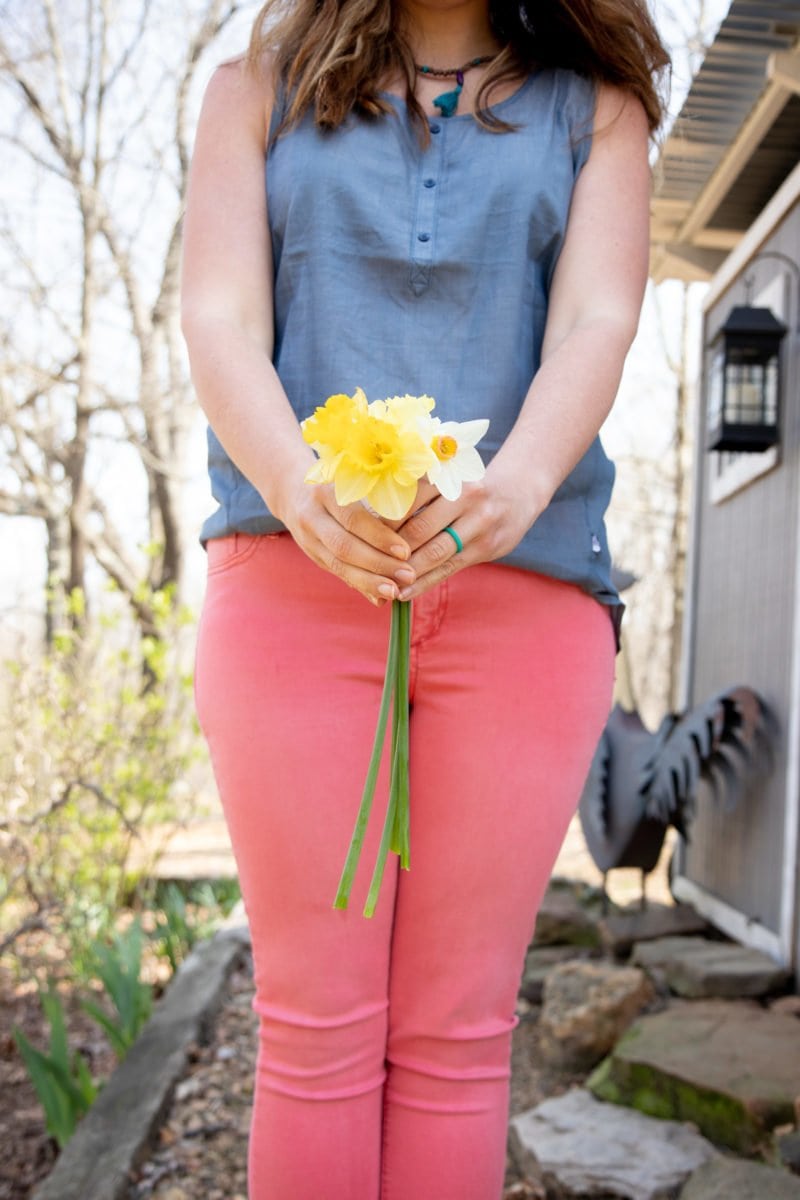 Shoulders-down shot of a woman in a blue shirt and pink pants, holding a bunch of daffodils