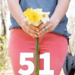 Shoulders-down shot of a woman in a blue shirt and pink pants, holding a bunch of daffodils, with a text overlay