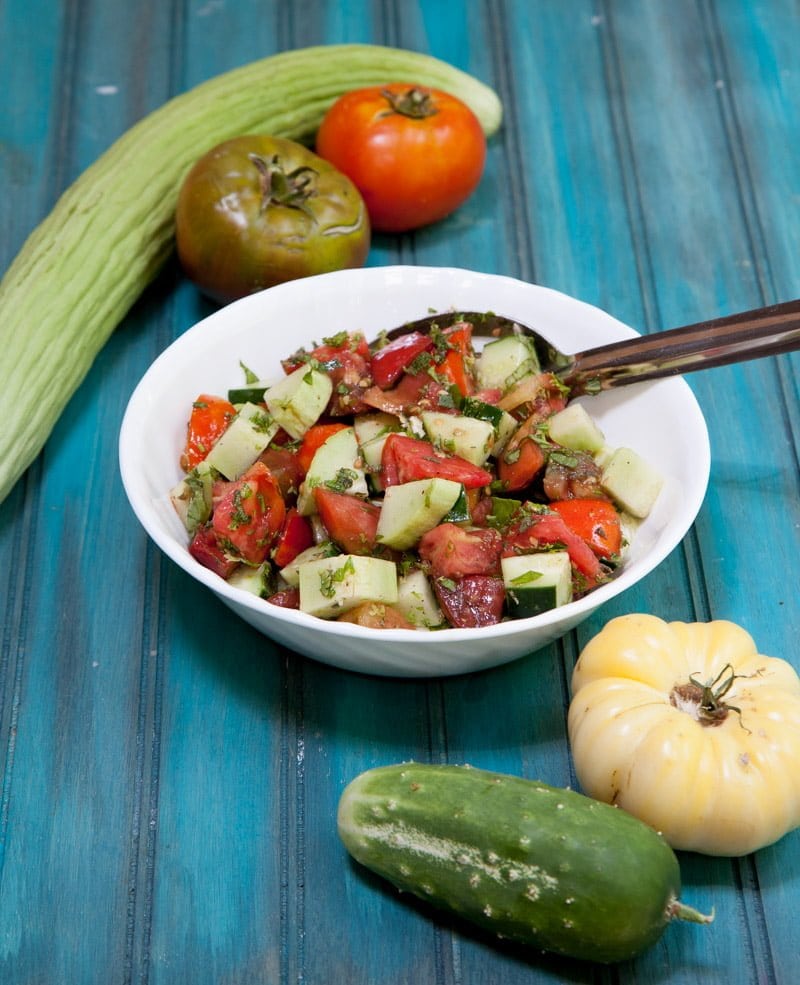 A spoon dips into a white bowl filled with herbed cucumber tomato salad