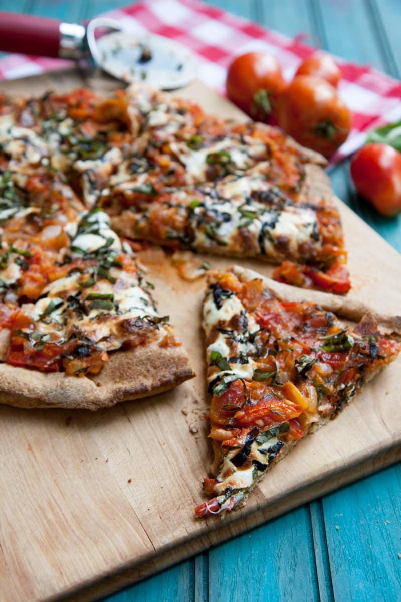 Close view of a slice of margherita pizza on a wooden cutting board with the rest of the pizza pie behind it.