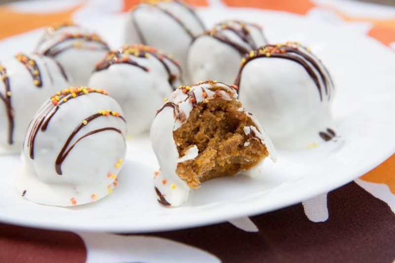 Pumpkin cake pops coated in white and drizzled with chocolate and orange sprinkles on a white plate. One cake ball has a bite taken out of it.