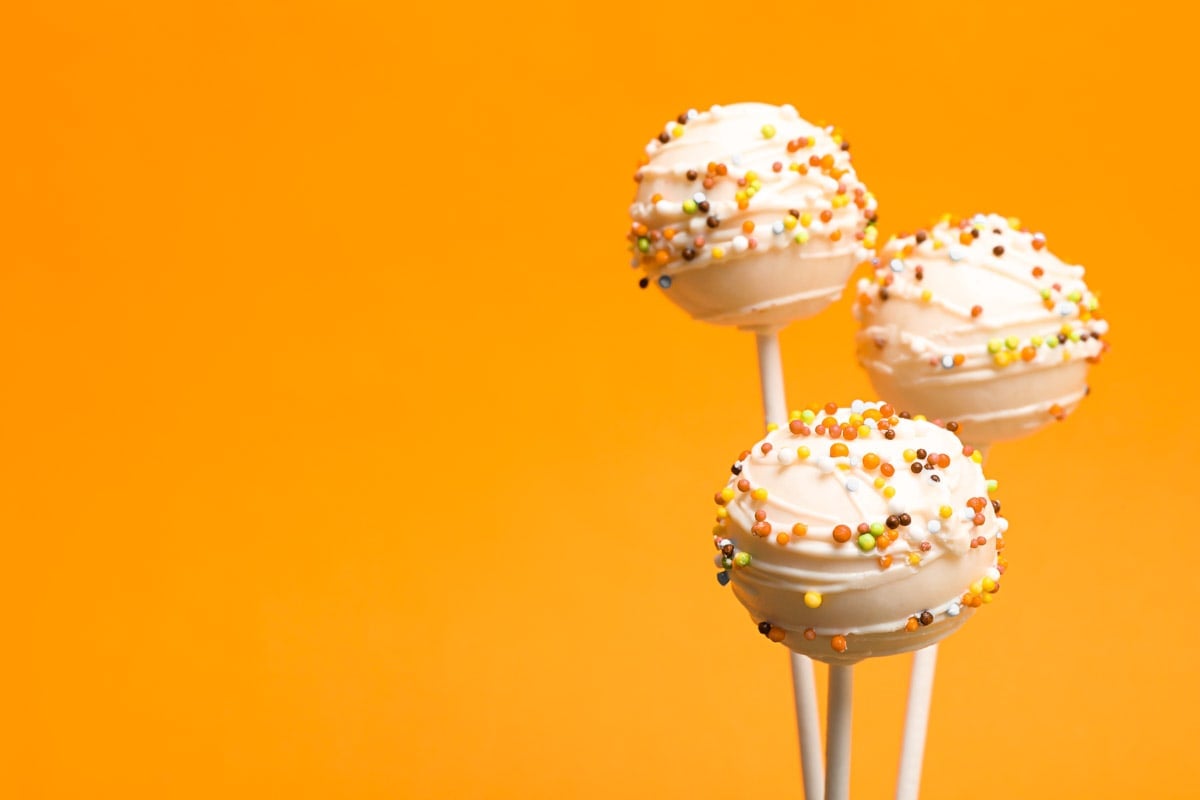 Three pumpkin cake pops in front of an orange background. The cake pops are covered in white candy melt and Halloween sprinkles.