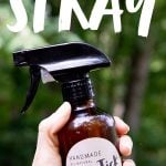 A hand holds an amber bottle of homemade all-natural tick and bug spray, with a text overlay that reads, "Homemade All-Natural Bug + Tick Spray".