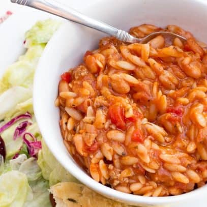 A bowl of Pasta e Fagioli sits on a white place, surrounded by lettuce and breadsticks.