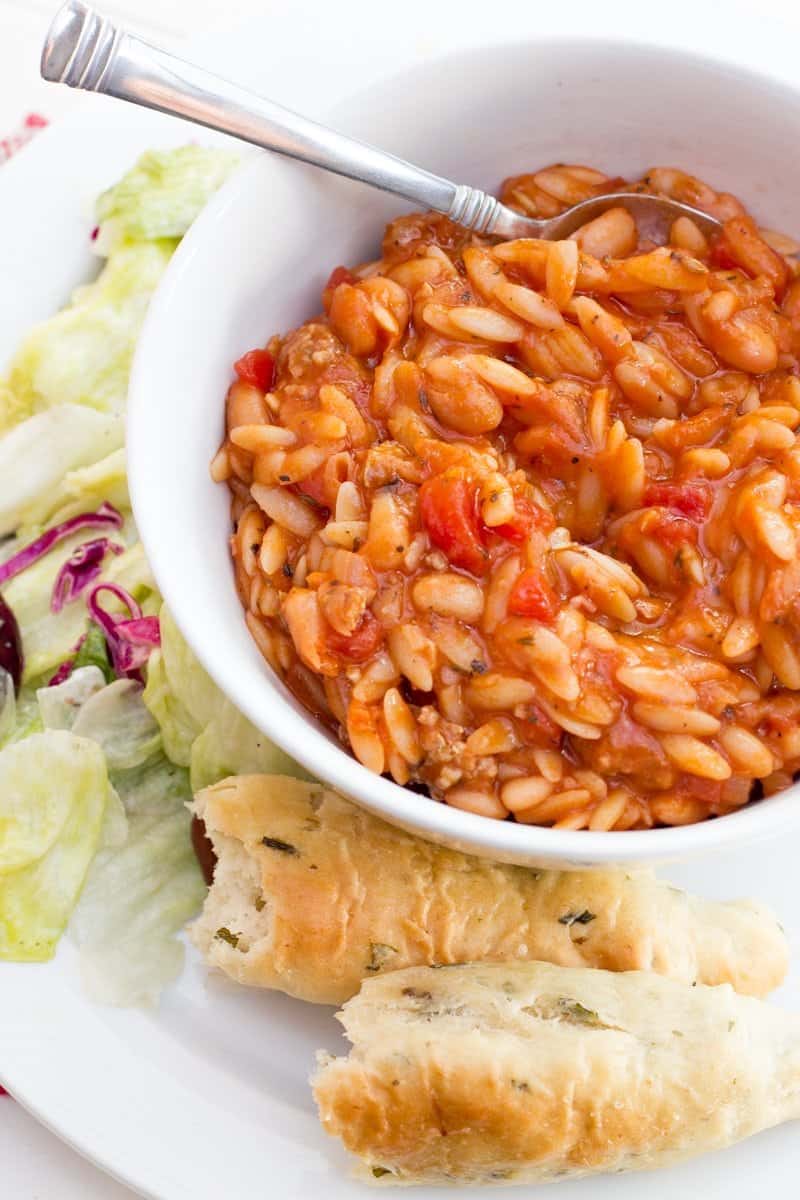 A bowl of Pasta e Fagioli sits on a white place, surrounded by lettuce and breadsticks.