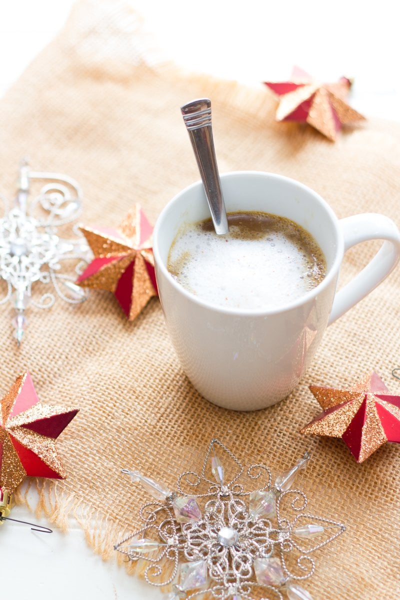 A homemade gingerbread latte in a white mug stands on a burlap mat amid holiday decorations.