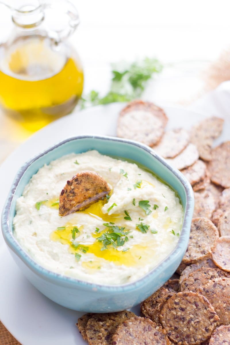 White bean dip drizzled with olive oil is in a light blue bowl, surrounded by crackers.
