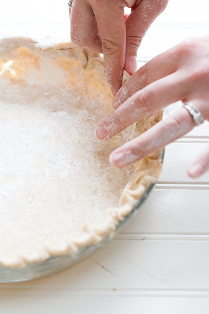 Two hands crimp pie dough around the edges of a pie plate.